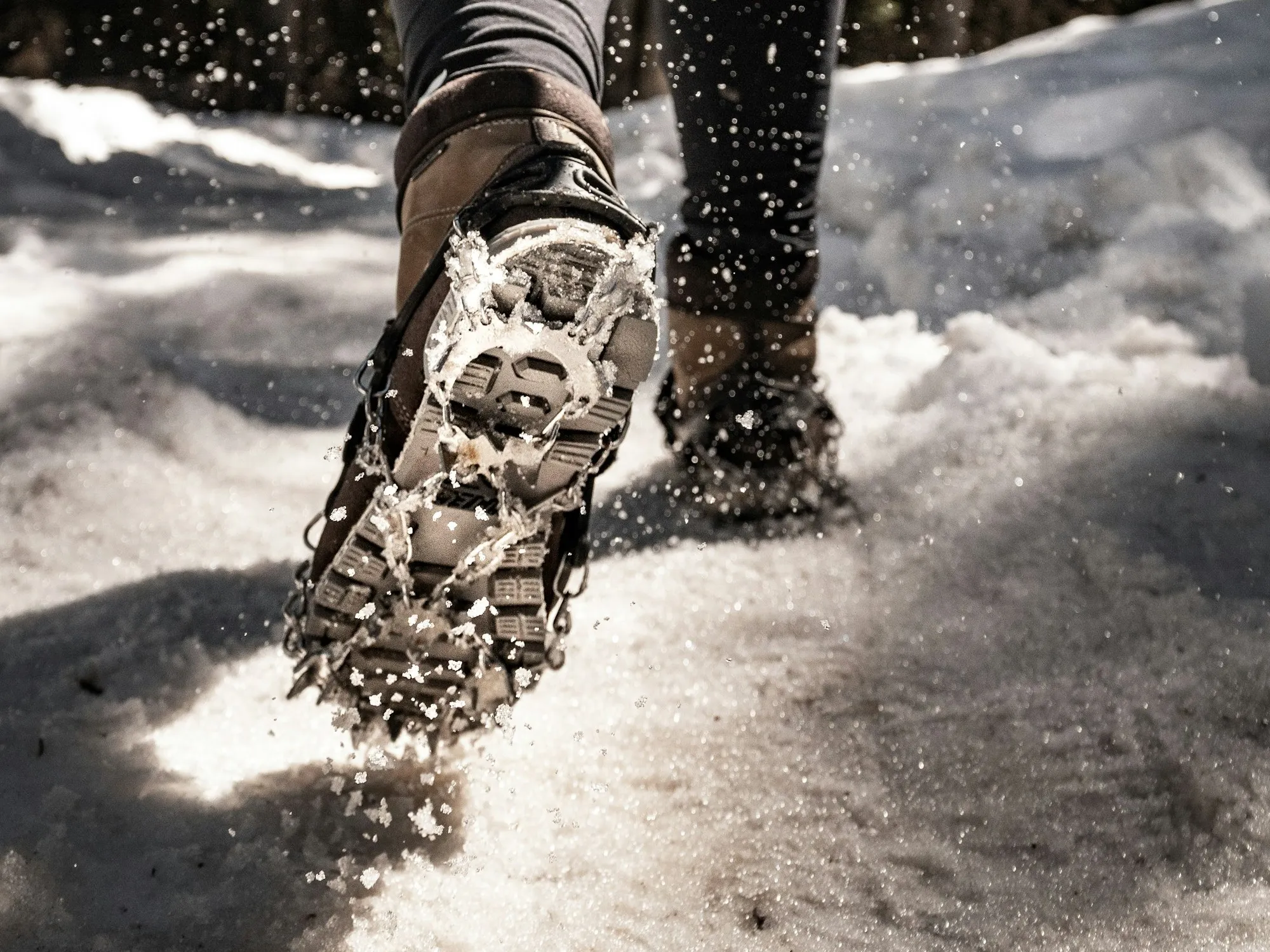 Close-up of a hiking boot fitted with a strap-on ice-traction cleat, chains and spikes across the sole, kicking up snow on a sunlit winter trail.