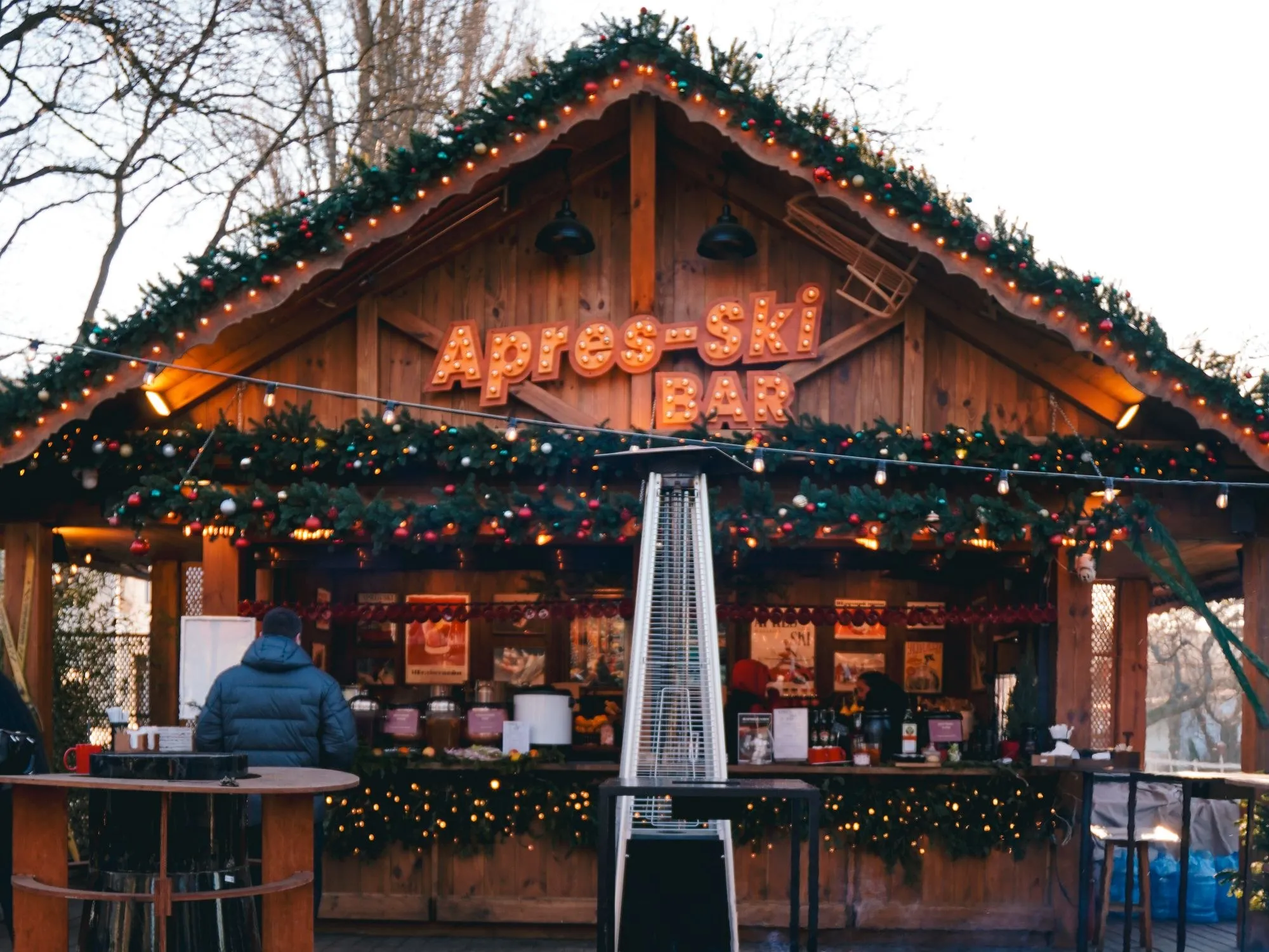An outdoor apres-ski bar at dusk with a lit 'Apres-Ski Bar' sign above a wooden chalet counter, garlands and fairy lights strung across the front, a bartender working the counter.
