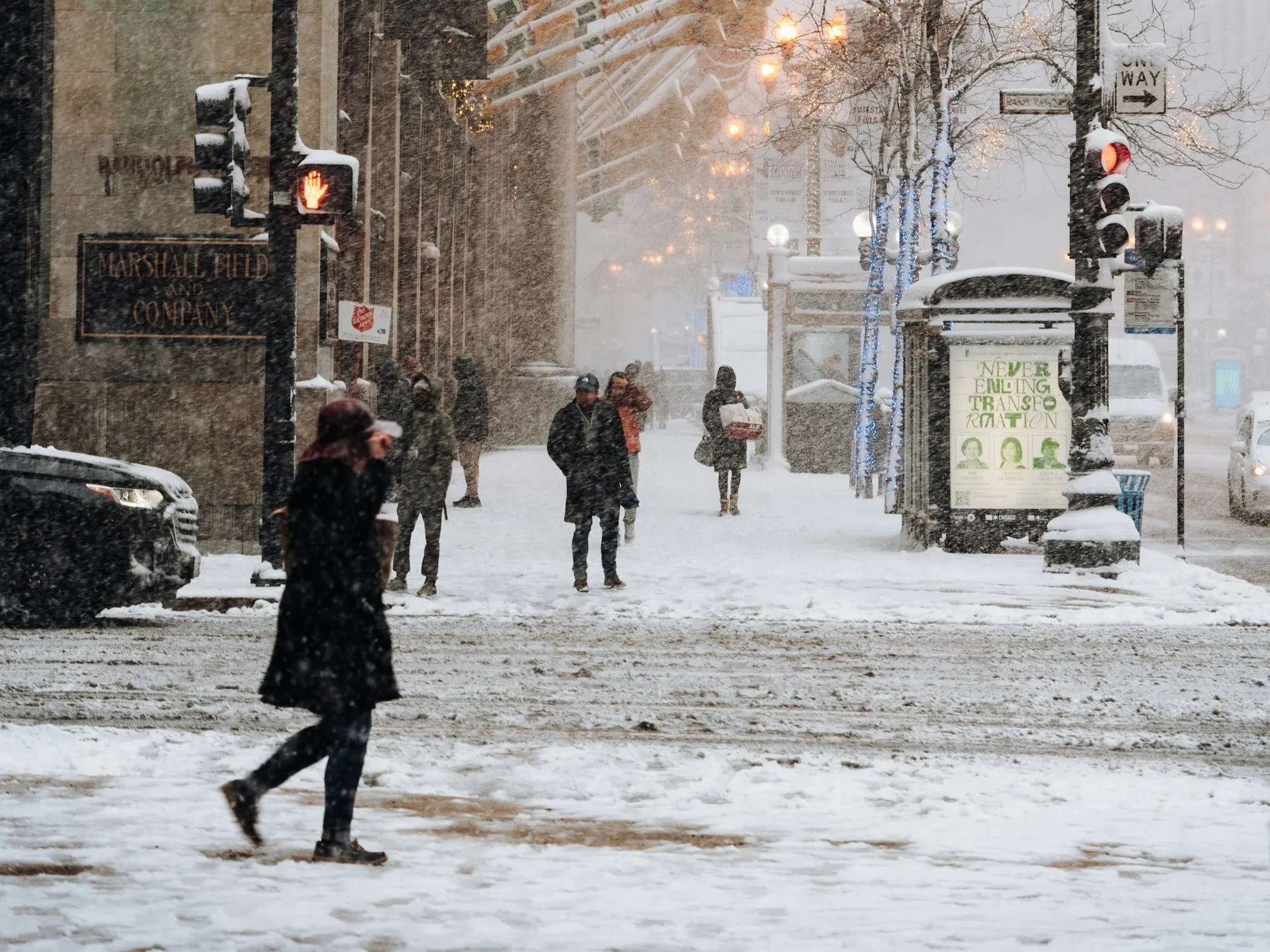 Pedestrians bundled in heavy winter coats crossing a snow-covered downtown Chicago street during a snowstorm, the historic Marshall Field and Company building behind them.