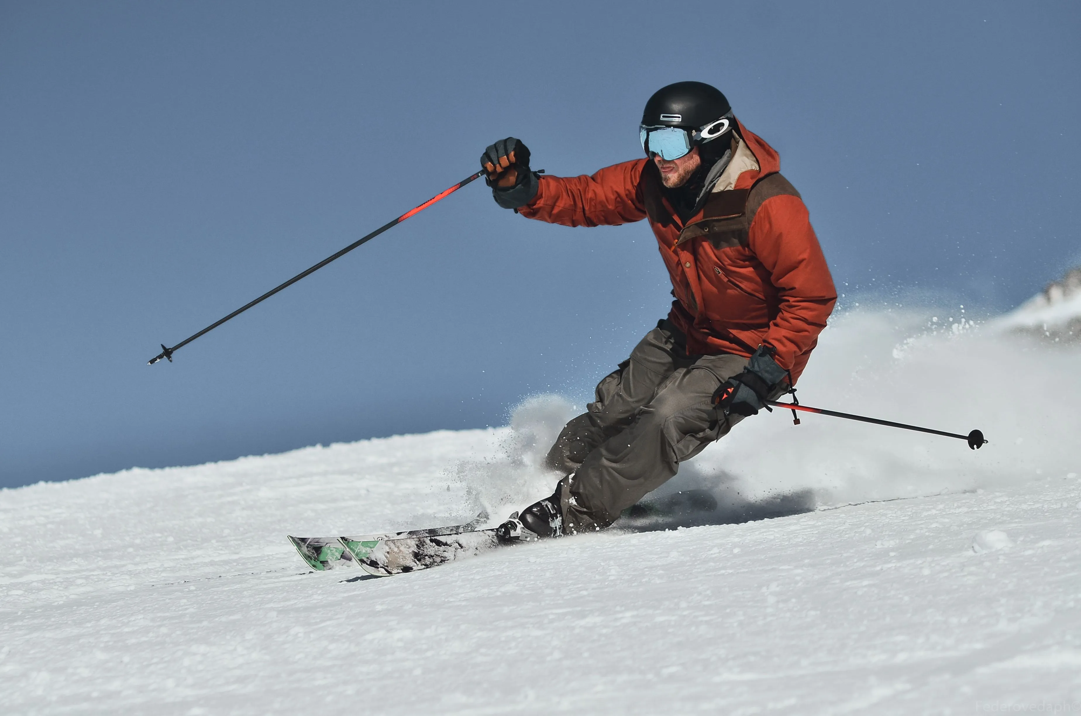 A skier in a red jacket and gray ski pants carving a turn through snow, spray flying, poles out, under blue sky.